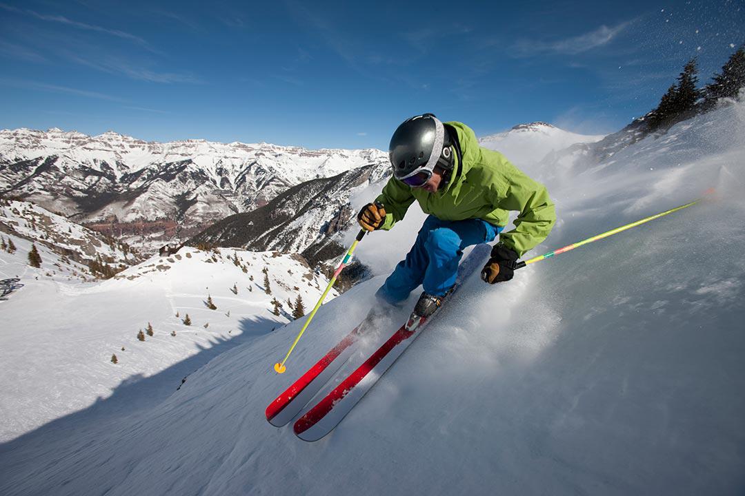 person wearing green jacket blue pants ski goggles black helmet skiing with red and white skis and yellow poles down a snowy slope with a brown and white mountain range in the background