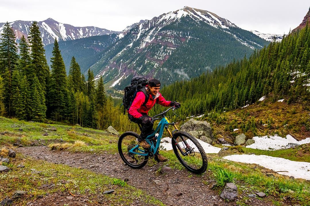 brunette woman wearing a red windbreaker and black backpack riding a blue bike on a dirt trail in Colorado in snow a forest of pine trees and snowy mountain peaks in the background