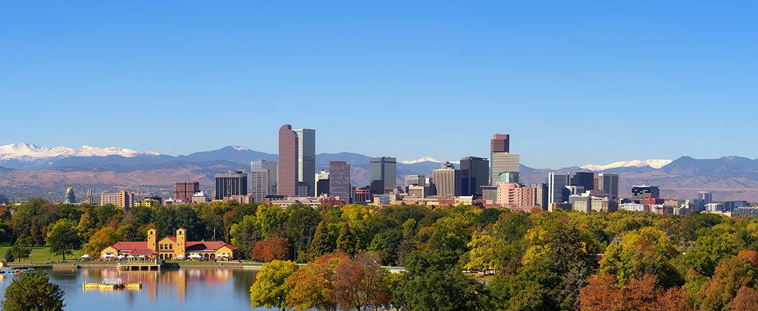 cityscape of Denver Colorado with dozens of buildings including skyscrapers with a mountain range in the background and trees and a lake in the foreground